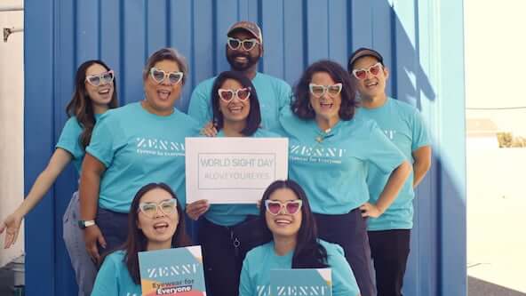Group wearing heart-shaped sunglasses. Signs read "ZENNI Eyewear for Everyone" and "WORLD SIGHT DAY #LOVEYOUREYES".