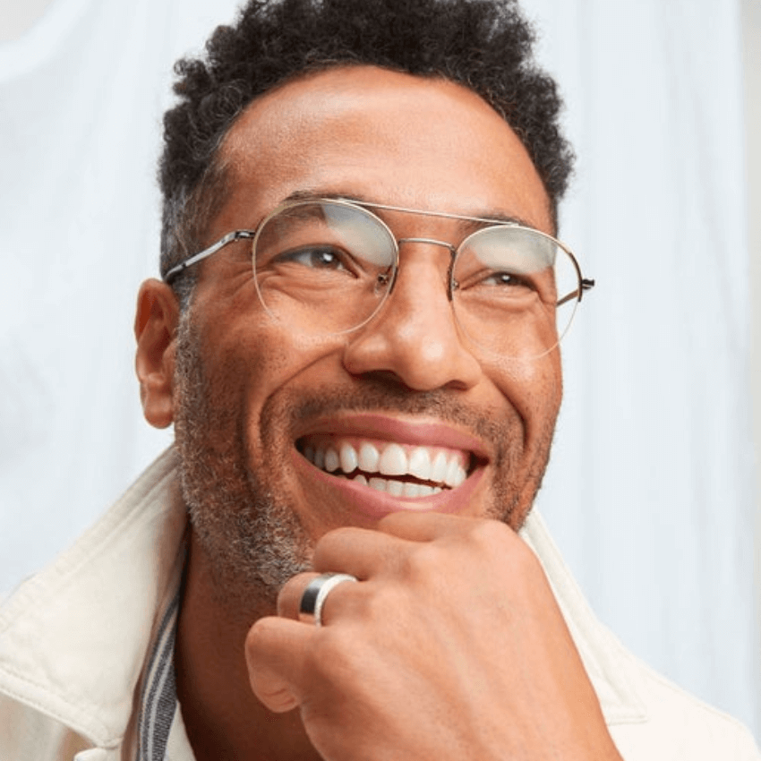 Man wearing thin-rimmed glasses and a white jacket, smiling, and holding his chin with a hand that features a silver ring.