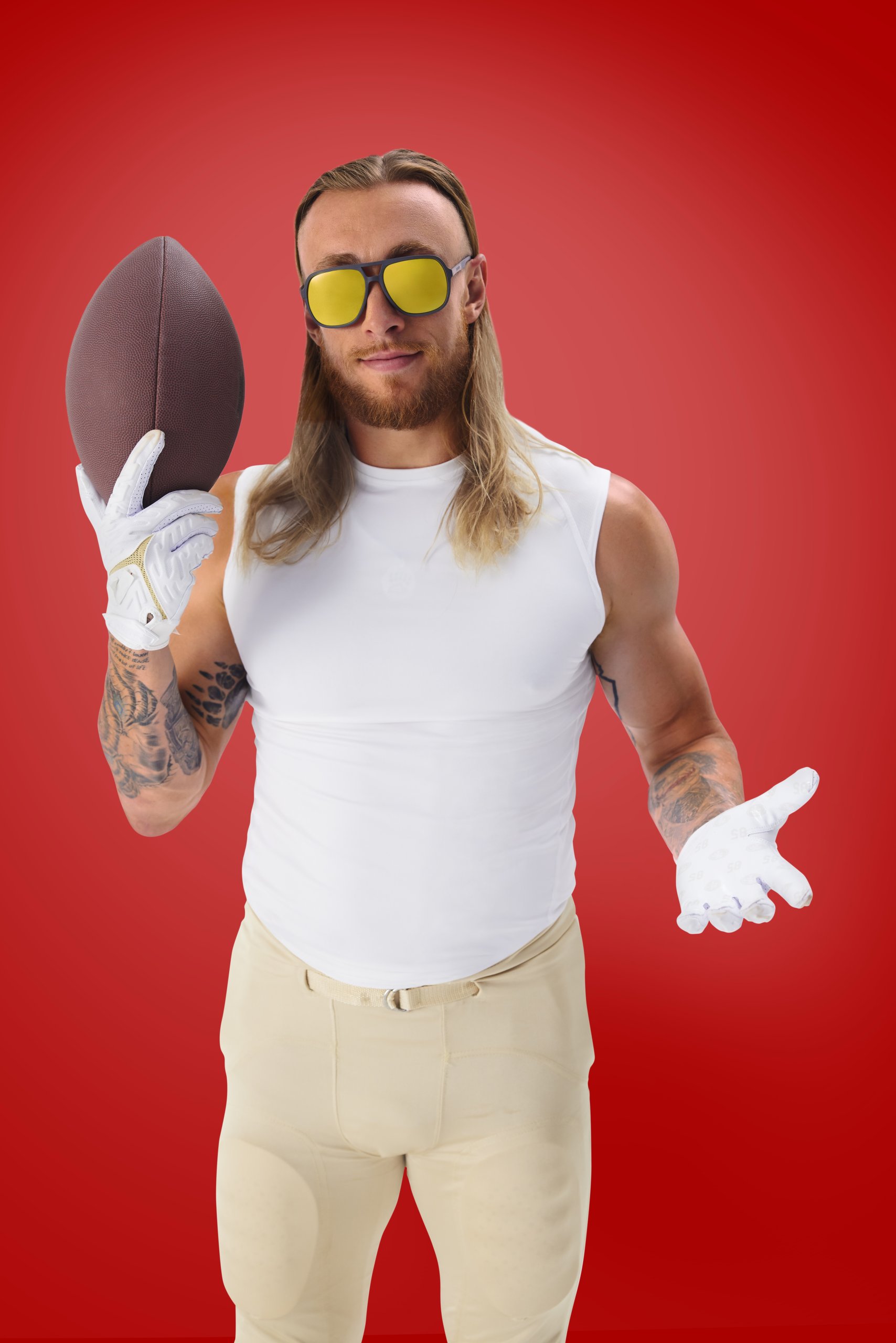 Man wearing yellow sunglasses, white gloves, white sleeveless shirt, and beige football pants, holding a football.