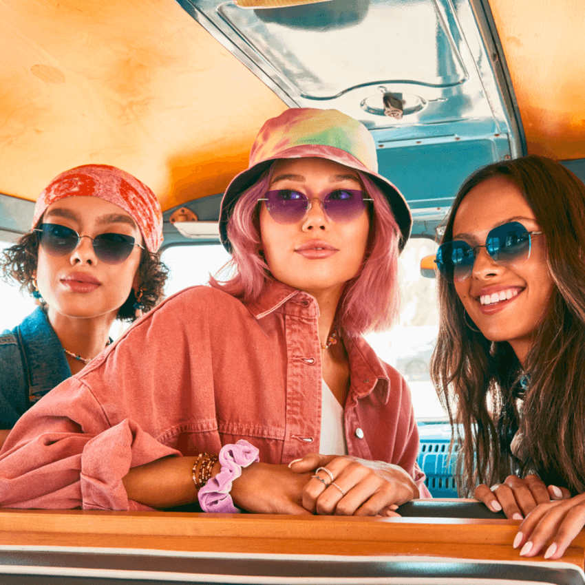 Three women wearing stylish sunglasses, vibrant headwear, and casual outfits inside a vehicle.