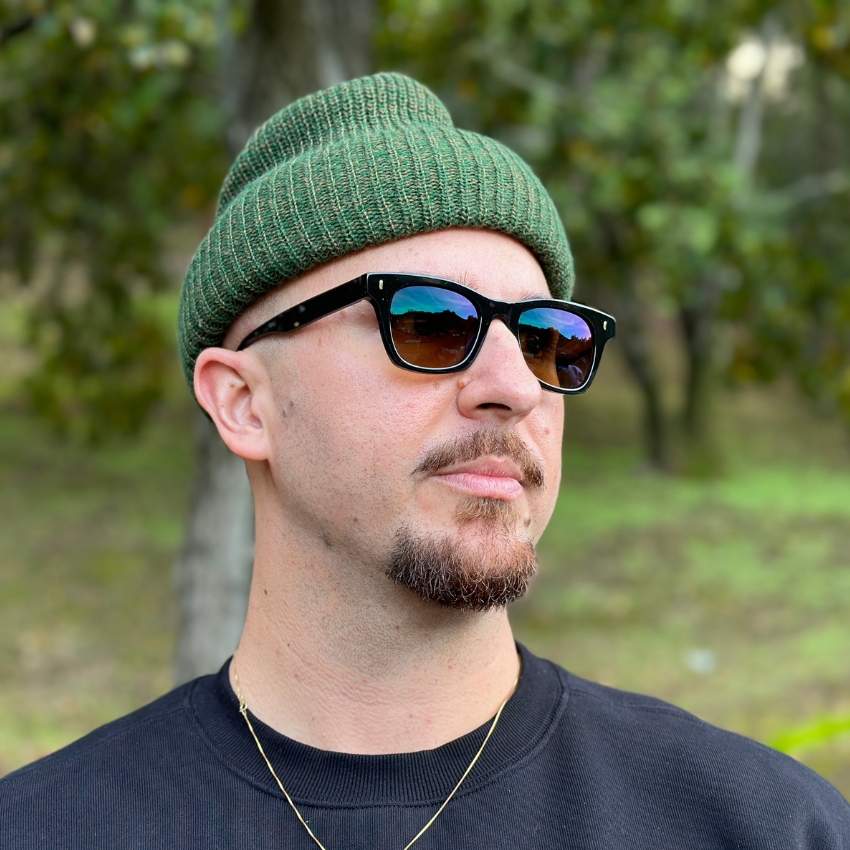 Man wearing black sunglasses with green knit beanie and black crew neck sweatshirt.