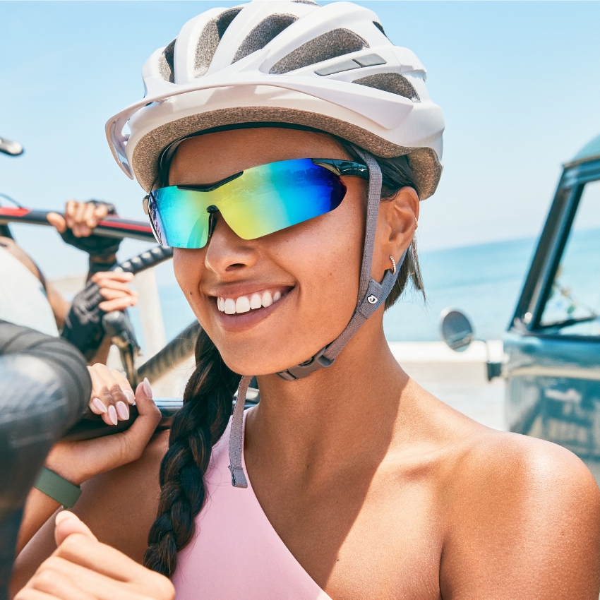 Woman wearing reflective cycling sunglasses and a white bike helmet, smiling outdoors.