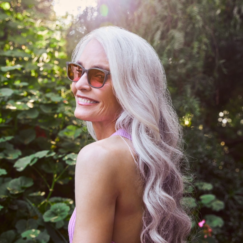 Woman with long silver hair wearing sunglasses and a sleeveless top, smiling while looking over her shoulder.