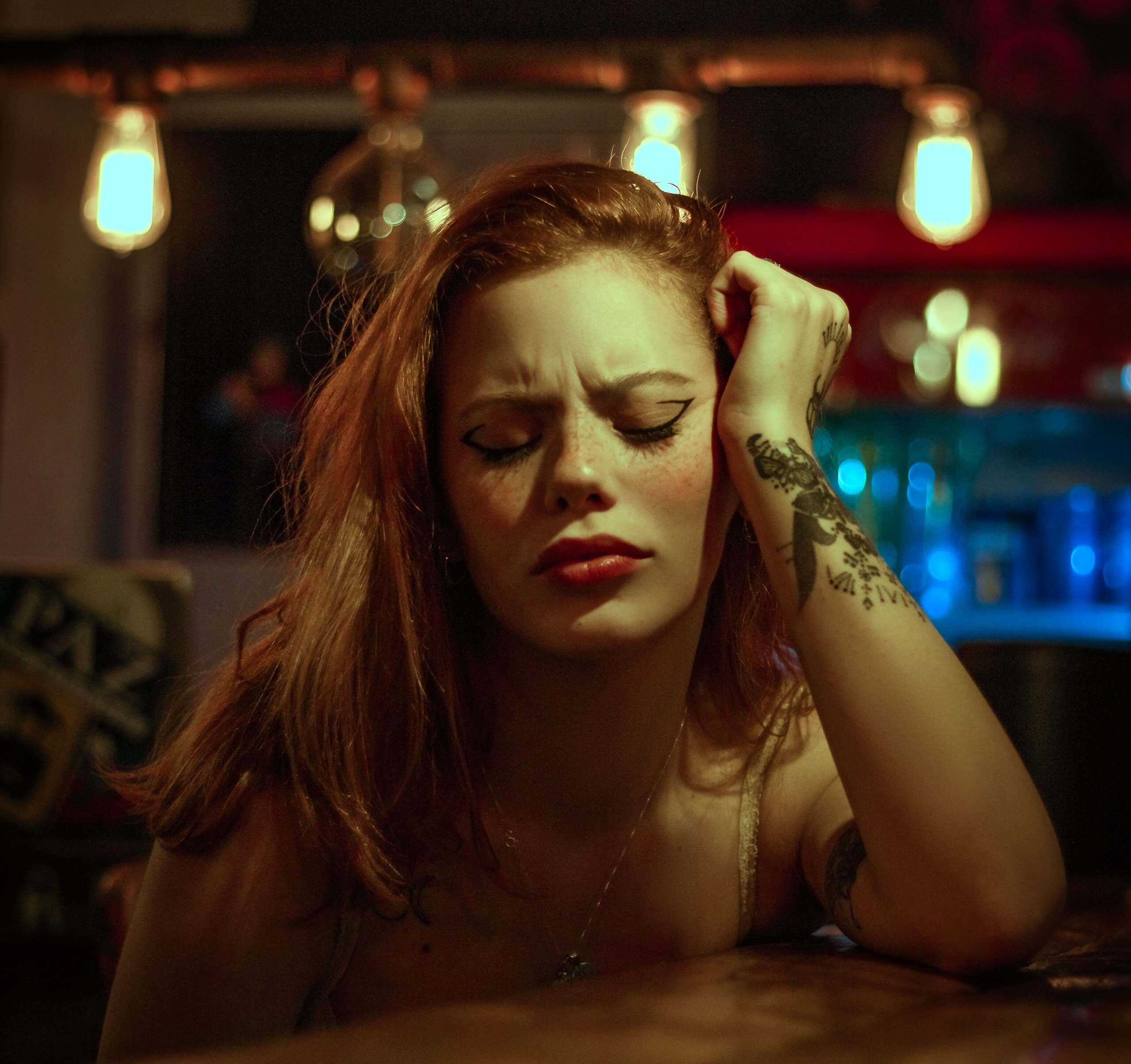 Woman with red hair and tattoos, wearing heavy eyeliner and red lipstick, resting her head on her hand.