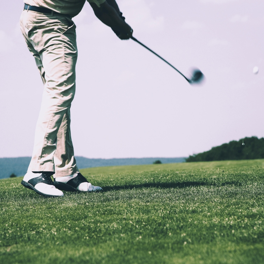 Golfer finishing a drive wearing white and black golf shoes and beige pants on a grassy course.