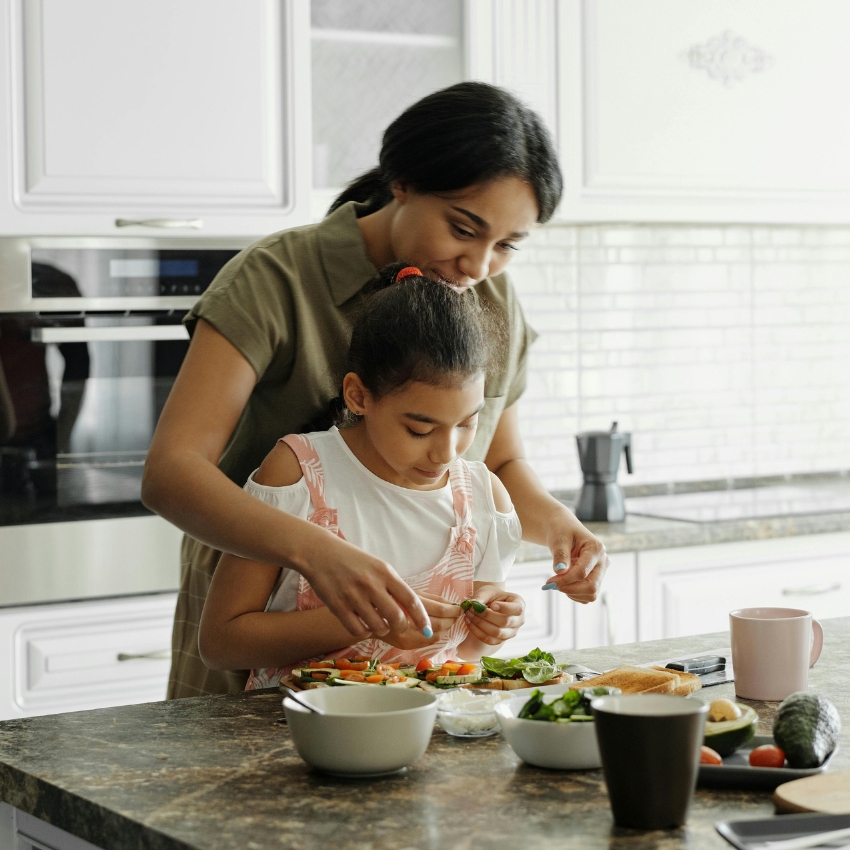 Mother helping daughter prepare food in a kitchen, with various bowls and ingredients on the counter.