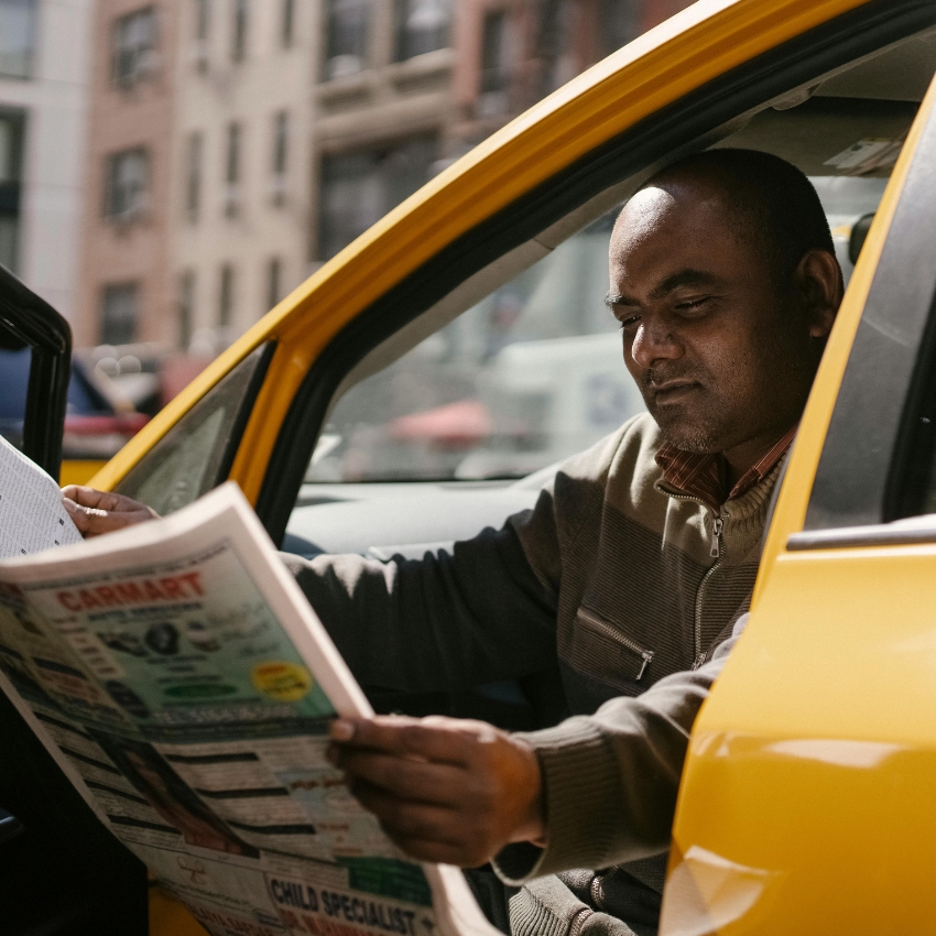 Man reading a newspaper in the driver's seat of a yellow vehicle. Visible text on newspaper: "CARMART"