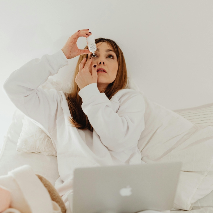 Woman applying eye drops while sitting in bed with a laptop in front of her.